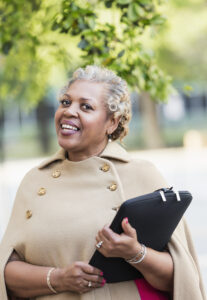 A mature African-American woman in her 50s with gray hair, standing outdoors, stylishly dressed in a beige coat. She is smiling at the camera.